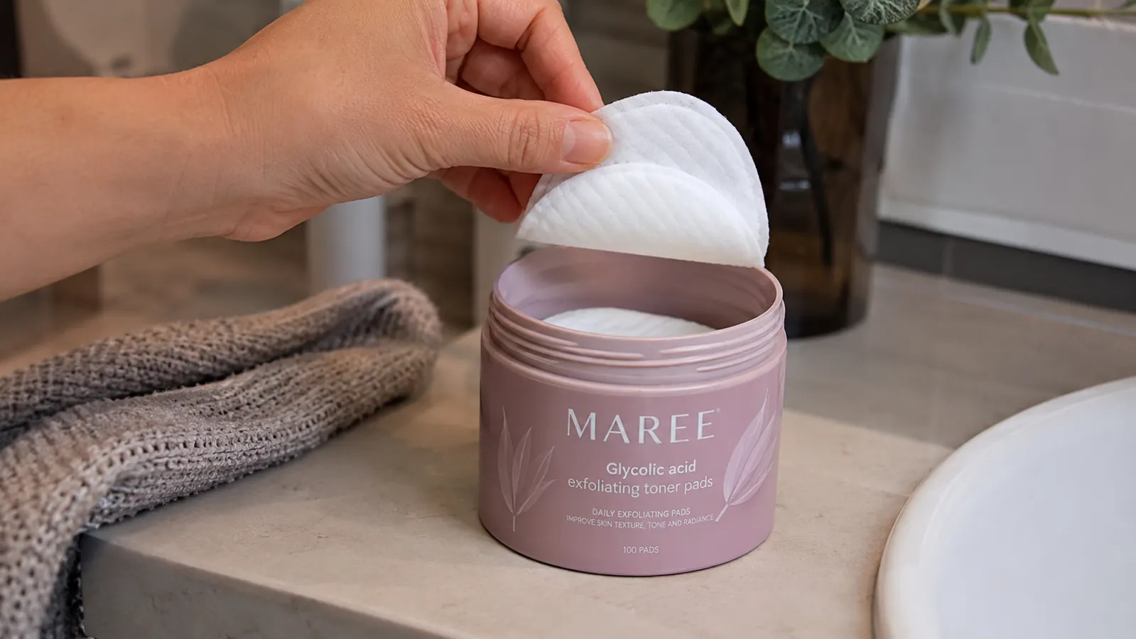 Close-up of a hand holding a MAREE glycolic acid pad above the open jar on a bathroom counter