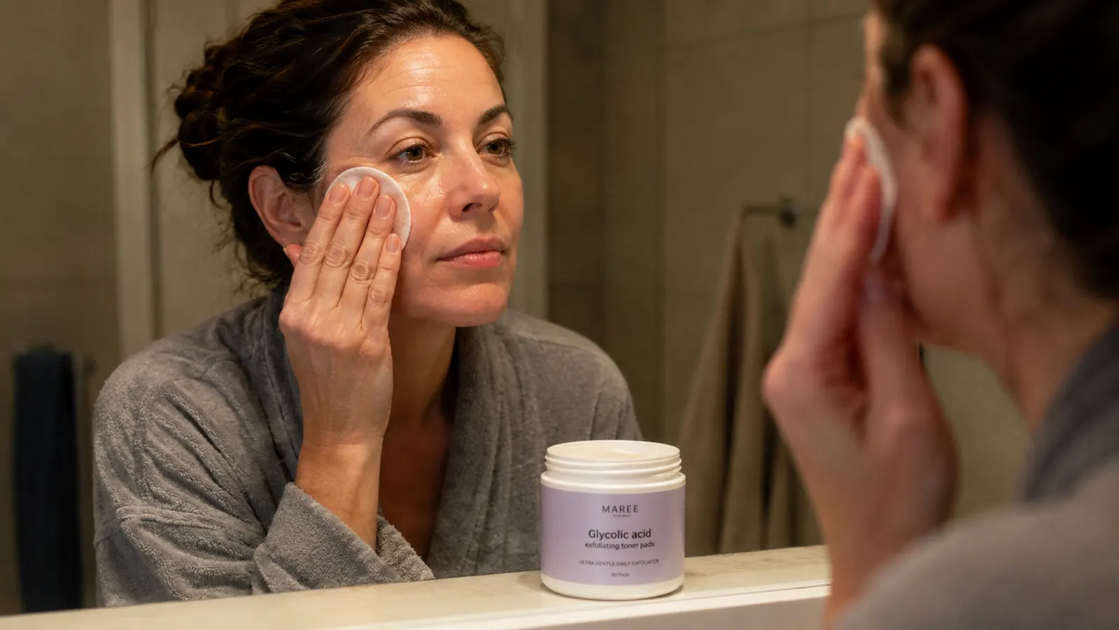 Hand wiping a MAREE glycolic acid pad across the cheek in a bathroom mirror after a long day