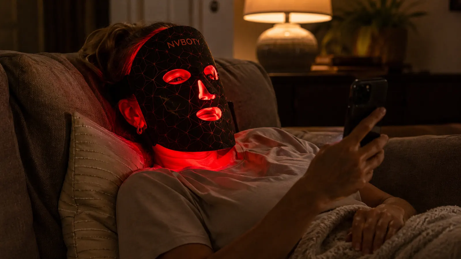 Woman wearing the NVBOTY red light mask hands-free while scrolling her phone on the couch