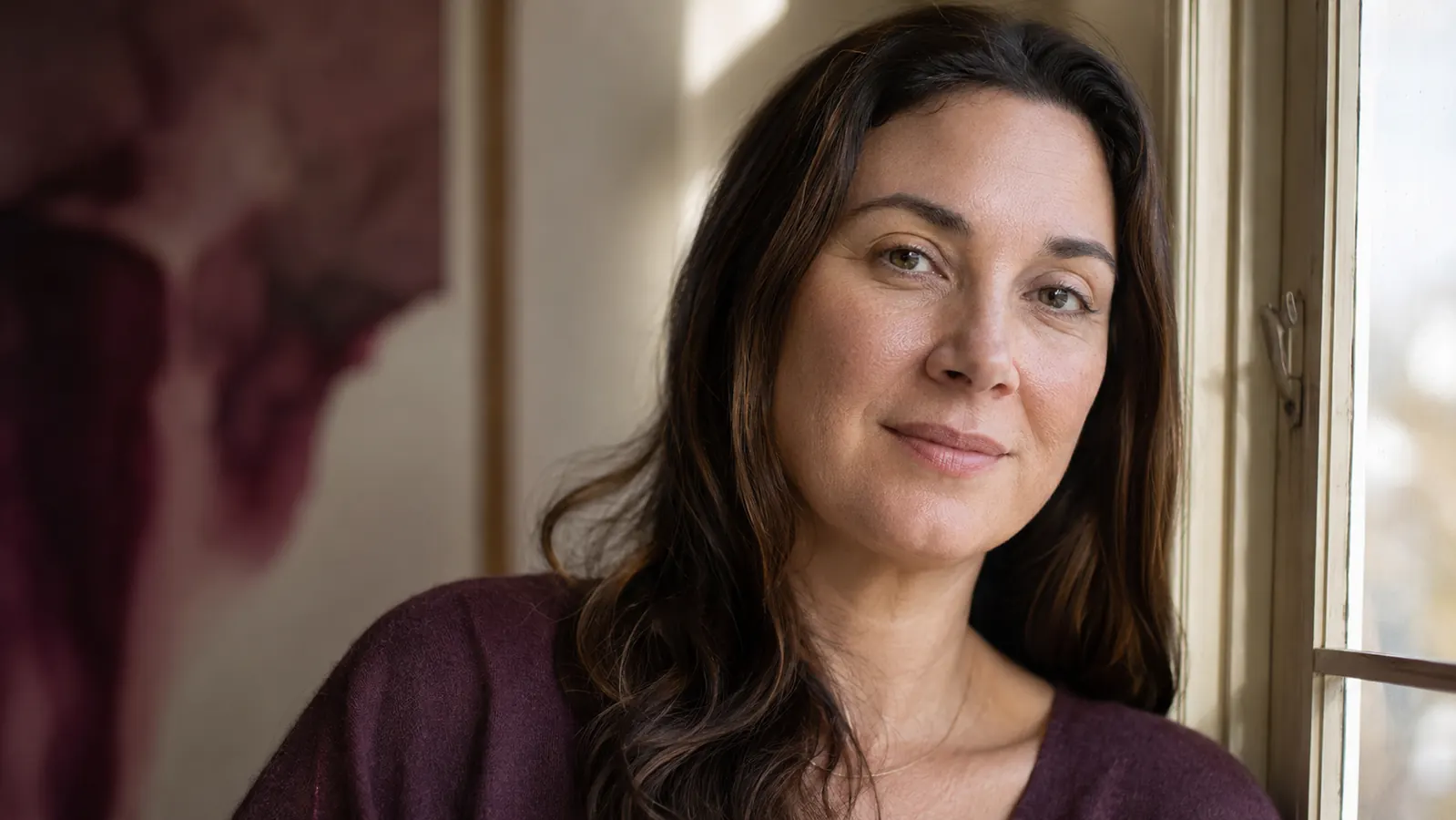 Woman with visibly calmer, more even-toned skin looking out a window in daylight, no makeup