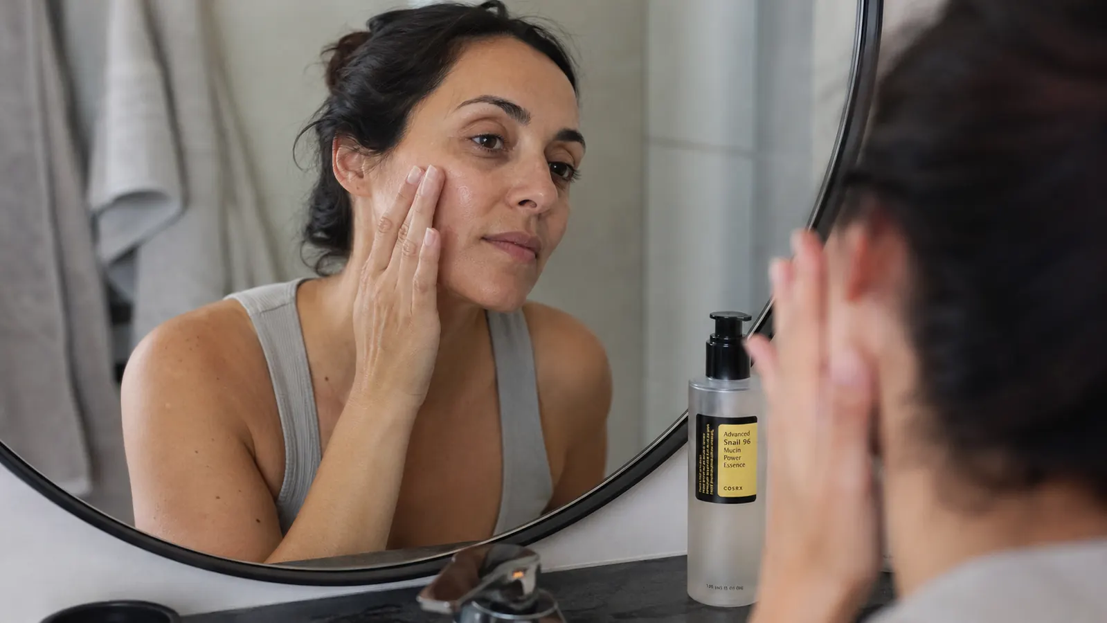 A hand pressing a few drops of snail mucin essence into windburned cheek skin at a bathroom sink